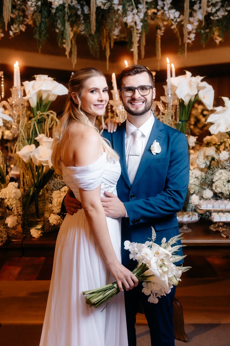 Alice e Daniel abraçados, com a noiva segurando o buquê de flores brancas e copos-de-leite ao fundo