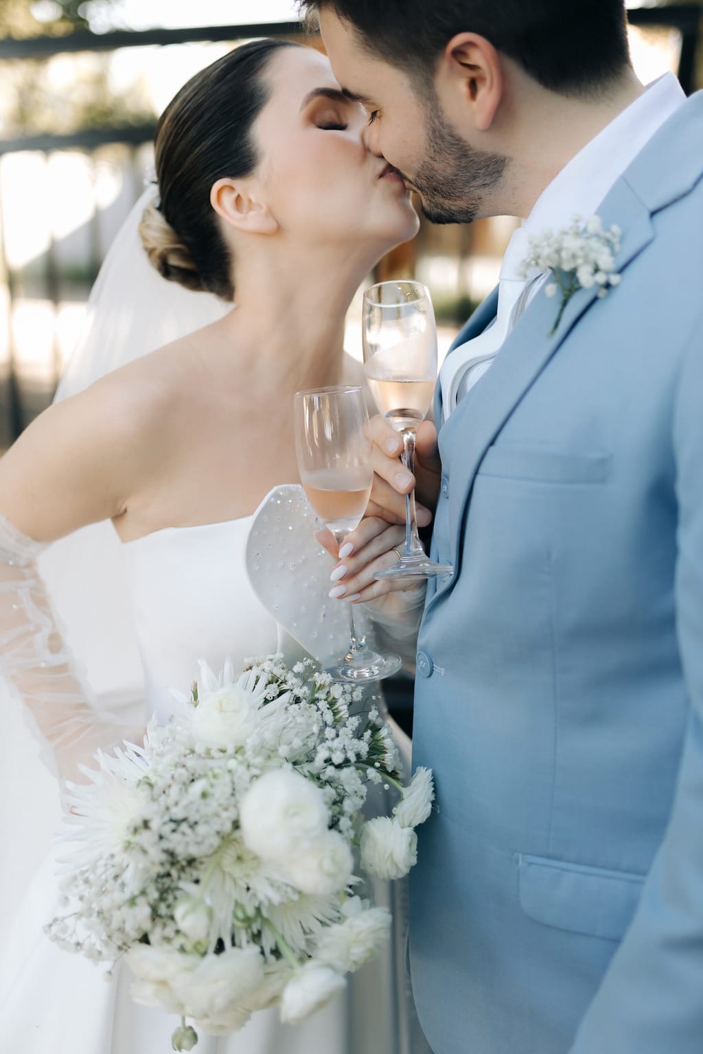Karoline e Angelo se beijando com taças de espumante, buquê de flores brancas