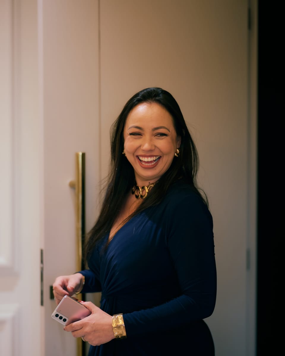 Michelle Nisimura rindo, de vestido azul-marinho e colar dourado, em um corredor de portas cor de creme