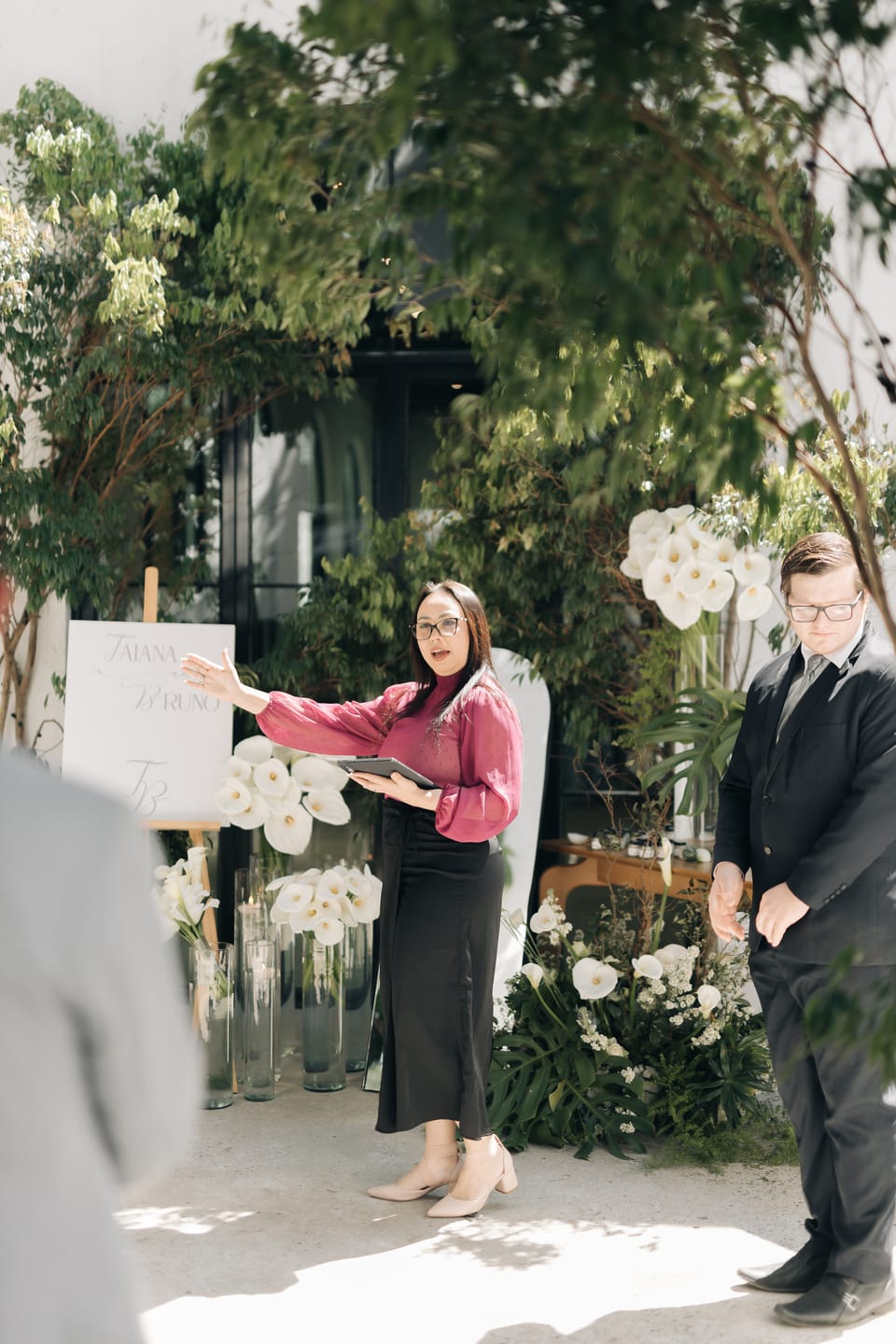 Michelle, de blusa magenta e prancheta na mão, coordena um assistente com um gesto do braço, ao lado da placa de boas-vindas e de arranjos de copos-de-leite